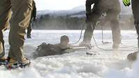 Eine Person ist im Eiswasser und wirft ihren Rucksack auf das Eis. Andere Personen beobachten das.