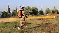 Ein Soldat trägt die Messvorrichtung waagerecht vor sich. Diese ist orange und etwa vier Meter lang.