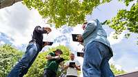 Fünf Personen stehen unter Bäumen im Kreis und bedienen je ein Tablet