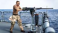 Auf einem Schiff auf See steht ein Soldat schießend hinter einem Maschinengewehr.