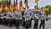 Soldaten marschieren auf einem Paradeplatz mit mehreren Truppenfahnen.