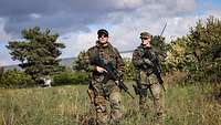 Zwei Soldaten gehen einen Feldweg entlang. Sie sind mit Gewehr, Schutzweste und Funkgerät ausgerüstet.
