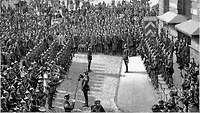 Eine Schwarz-Weiß-Zeichnung von Soldaten mit Pickelhaube, die auf einem Marktplatz angetreten sind