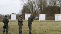 Drei Soldaten stehen auf einer Wiese. In der Ferne stehen Zielscheiben. Ein Soldat zielt mit seiner Waffe auf diese.