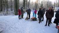 Eine Gruppe Menschen steht in einem Winderwald an einer Feuerstelle. 