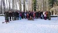 Eine Personengruppe steht in einer verschneiten Landschaft um ein Lagerfeuer zusammen.