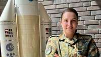 Oberleutnant Kerstin ist Weltraumlageoffizier. Sie steht in grüner Uniform in einem Gebäude vor einem Raketenmodell.