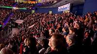 Viele Menschen sitzen auf Tribünen in einer Arena. 