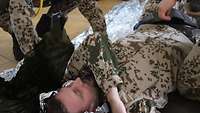 Ein Soldat liegt am Boden, er spielt die verletzte Person und wird von weiteren Soldaten in eine Rettungsdecke gelegt