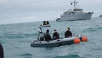 Mehrere Personen sitzen auf zwei Schlauchbooten, im Hintergrund ein graues Schiff auf hoher See