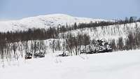 In einem bergigen Gebiet voller Schnee fahren Soldaten auf Kettenfahrzeugen einen Hang hinunter