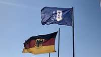 Die NATO-Flagge, die KFOR-Flagge und die Bundesdienstflagge wehen vor blauem Himmel. 