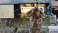 Ein Soldat in Flecktarnuniform steht vor einer Geländekarte, die hinter ihm hängt.