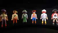 Sechs verschiedene Playmobilfiguren stehen vor einer schwarzen Leinwand.
