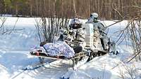 Ein Skidoo fährt mit Anhänger durch Schnee. Darauf sitzt ein Soldat mit einer großen Waffe.