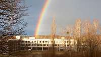 Im Hindergrund eines Gebäudes und eines Waldstückes ist am Horizont ein Regenborgen zu sehen 