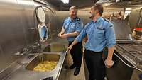 Zwei Marineangehörige in Uniform beim Kartoffelschälen in der Schiffsküche.