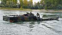 Ein Motorboot auf dem ein Soldat steht schwimmt im Wasser und schiebt eine Schwimmbrücke vor sich her