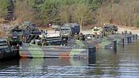 Mehrere Motorboote fixieren eine Faltschwimmbrücke mit militärischen Fahrzeuge darauf