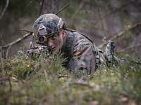 Ein Soldat liegt im Wald hinter einem Baum mit der Waffe im Anschlag.