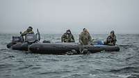 Mehrere Soldaten sitzen in einem Schlauchboot was im Wasser schwimmt, Taucher halten sich daran fest