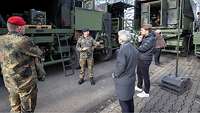 Eine Gruppe Menschen steht vor einem Soldaten zwischen mehreren Fahrzeugen und hört im zu.