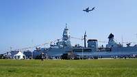 A ship of the German Navy lying in the port. A transport aircraft of the German Air Force hovers above it.