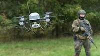 Eine Drohne vom Typ MIKADO im Flug als Portraitaufnahme. ImHintergund steht ein Soldat.