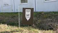 Ein schwarzes Schild mit der Aufschrift TCP-MP steht vor einem Container. 