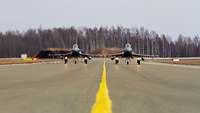 Zwei Kampfjets Starten zum Flug auf einem Rollfeld im Hintergrund sind Bäume zusehen