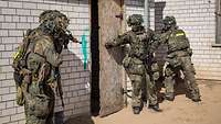 Drei Soldaten im Kampfanzug stehen vor einem Gebäude bereit, um eine Tür zu öffnen