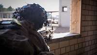 Ein Soldat zielt mit einem auf eine Festeröffnung aufgelegten Maschinengewehr im Anschlag aus einem Gebäude heraus