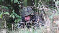 Ein Soldat liegt neben einem Baum im Grad. Er hat sein Gewehr in Anschlag.