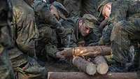 Mehrere Soldaten knien auf dem Boden und bauen ein Gestell aus Holzstämmen