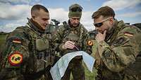 Drei Soldaten schauen auf einem Truppenübungsplatz auf eine taktische Karte und sprechen miteinander