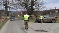 Ein Soldat stoppt ein Militärfahrzeug mit einer Winkerkelle. Rechts von ihm steht ein weiterer Soldat vor einem Fahrzeug.