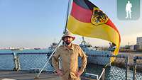 Ein Soldat steht in Uniform vor der Heckflagge eines Schiffes