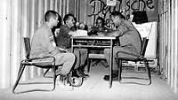 Schwarz-Weiß-Foto von vier Soldaten, die an einem Tisch sitzen. Ein Soldat hält ein Mikrofon