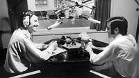 Schwarz-Weiß-Bild von zwei Männern, die an einem Tisch mit Mikrofonen sitzen 