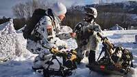 Ein Soldat wird von einem anderen Soldaten interviewt. Beide tragen Schneetarn und knien im Schnee. 