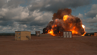 Eine Feuerwolke breitet sich über einem Feld aus. Ein Gebäude steht davor.