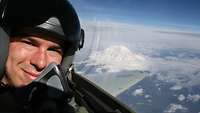 Ein Pilot mit Helm sitzt in einem Flugzeug. Im Hintergrund ist ein schneebedeckter Berg zu sehen.