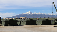Vor einer Bergkulisse stehen grüne Bundeswehrzelte, im größeren Abstand dahinter große beige Zelte 