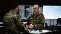 Zwei Soldaten in Uniform stehen sich im Gespräch gegenüber, im Hintergrund sind Computerbildschirme. 
