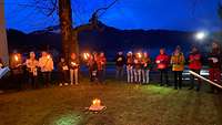 Menschen stehen mit Fackeln in der Hand in einer Reihe auf einer Wiese vor drei brennenden Kerzen.