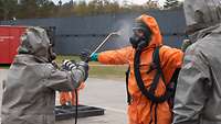 Mensch im grauen Schutzanzug spritzt eine Person im orangenen Overall mit Atemschutzmaske ab.