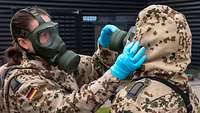 Eine Soldatin hilft der anderen beim Anlegen der Atemschutzmaske. Beide tragen Schutzanzüge.