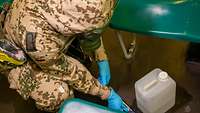 Eine Soldatin entnimmt in Schutzkleidung, Atemschutzmaske und Schutzhandschuhen eine Probe