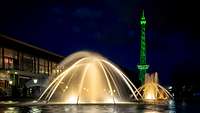 Ein mit Licht angestrahlter Springbrunnen und Metallturm, daneben ein Gebäude mit großen Fenstern