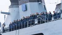 Auf einem Seitendeck eines grauen Schiffs stehen Marinesoldaten in blauer Uniform-Arbeitsjacke